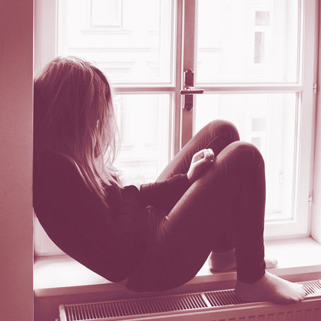 A person in dark clothing sits on a windowsill with knees up, looking contemplative. The room is softly lit with a pinkish hue.