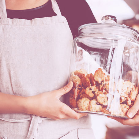 Person wearing an apron holds a glass jar filled with cookies. Blurred background with warm lighting suggests a cozy bakery setting.