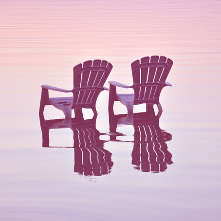 Two empty Adirondack chairs sit partially submerged in calm water, reflecting a serene pink and blue sunset sky.