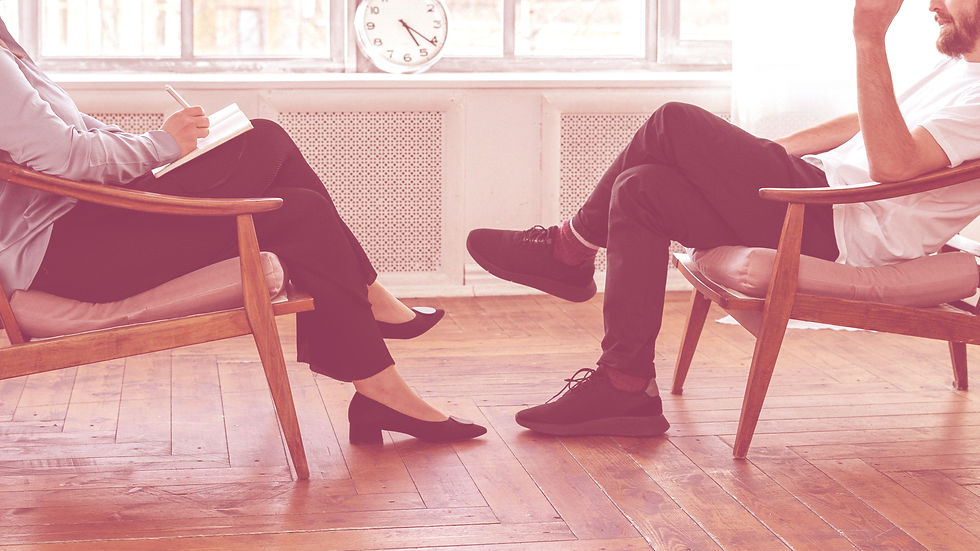 Two people sit in armchairs on a wooden floor, engaging in conversation. One person writes notes. A clock and window are in the background.