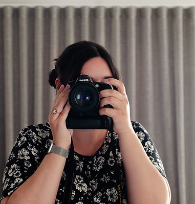 Ellie capturing her own portrait with her DSLR in a mirror. Her face is covered mostly as she looks through the lens.