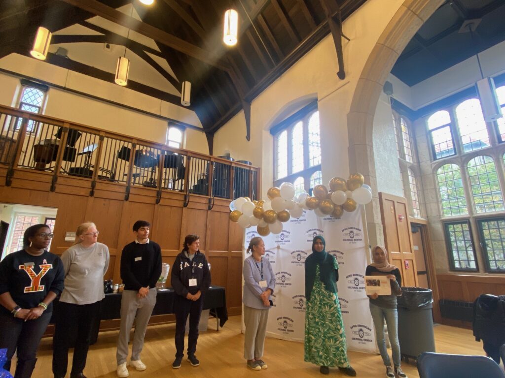 Seven people stand in a line watching one of them display a document