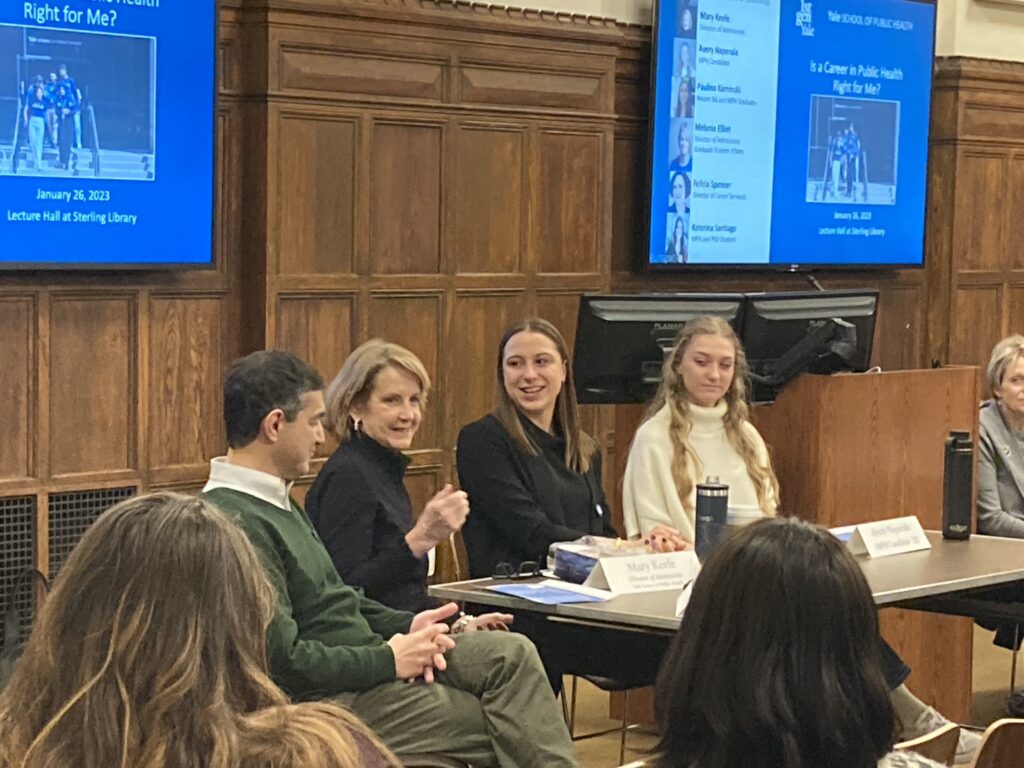Four people seated at a table with one person gesturing and talking to an audience