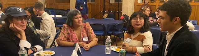 Four people seated at a table with food and beverages talk.