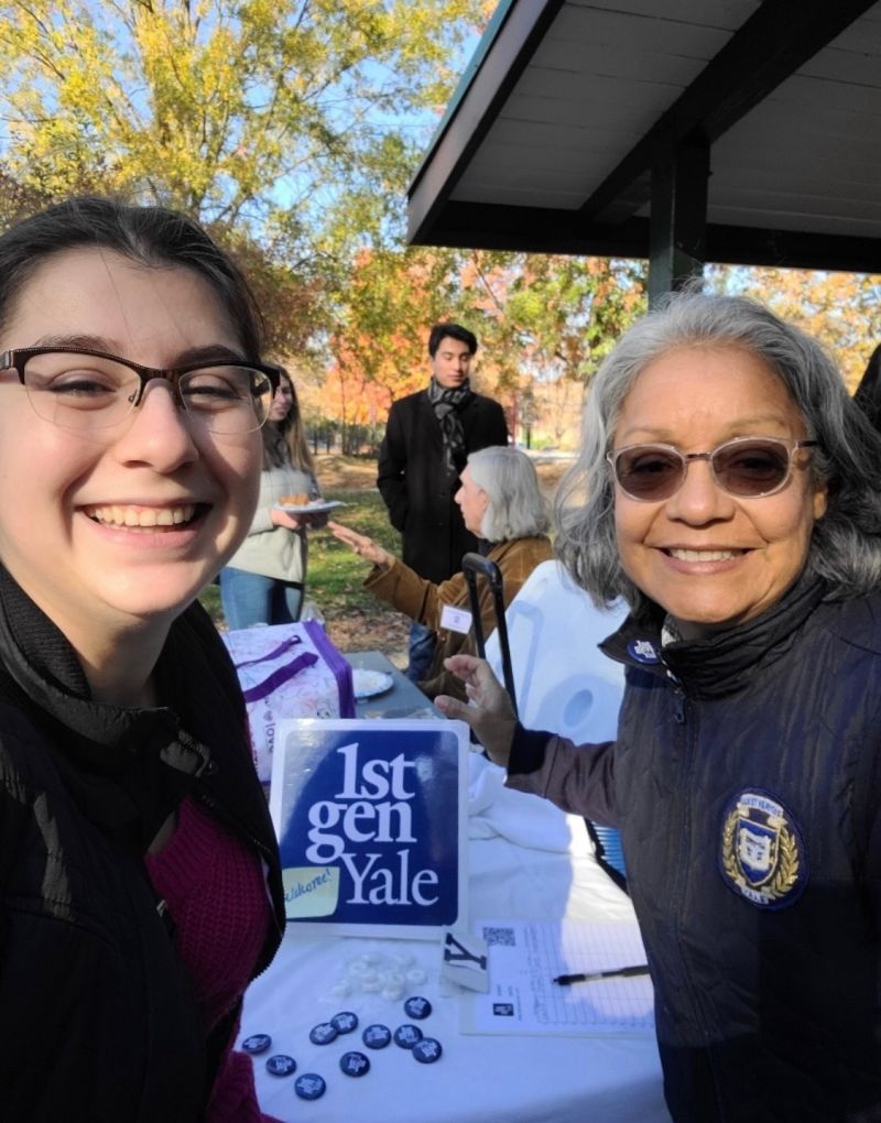 Two women standing at a table outside with a 1stGenYale sign