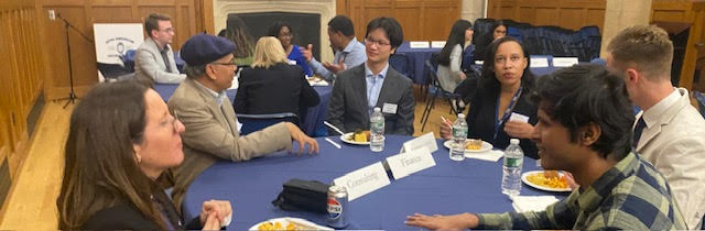Six people seated at a table talk. There are several cards with career fields on them, and another table of people in the background.