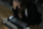 A person sits at a desk with a laptop, resting their head in one hand and looking down. A cup, phone, and glasses are nearby, suggesting they may be feeling stressed or overwhelmed.
