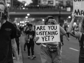 A person holds a large sign reading “ARE YOU LISTENING YET?” while standing in a crowd during a protest. The photo is black and white.