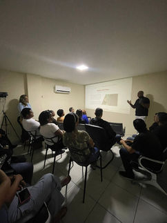 Um homem e uma mulher apresentam, por meio de uma imagem projetada em uma parede branca, uma imagem de um website que mostra um vídeo de um escritório e um bloco de texto institucional para um grupo de pessoas sentadas em cadeiras. 