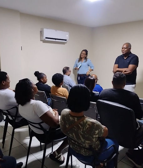 Um homem e uma mulher fazem uma palestra em frente a um grupo de pessoas sentadas em cadeiras dentro de uma sala iluminada.
