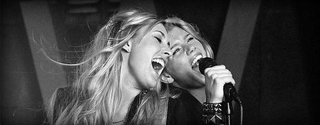 Primo piano di due belle ragazze bionde che cantano a piena gola, sorridenti e felici.
