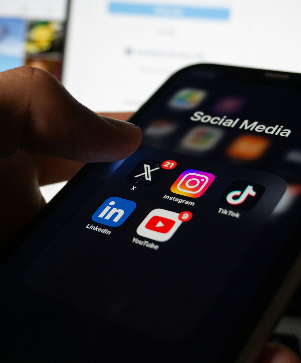 Hand holding a smartphone displaying social media apps: X, Instagram, LinkedIn, TikTok, YouTube. Notifications visible on X and YouTube.