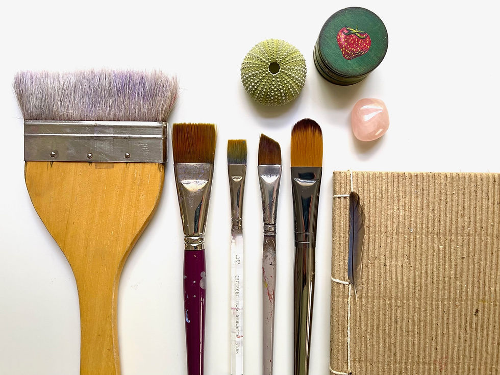 A collection of art supplies including paintbrushes, a handmade journal, a seashell, and a rose quartz crystal arranged neatly on a white background.