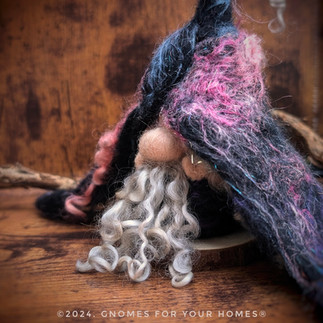 "Close-up of a needle-felted gnome with a beige felted face and long curly white beard, wearing a black hood decorated with pink and blue wispy wool accents. © 2024, GNOMES FOR YOUR HOMES®"