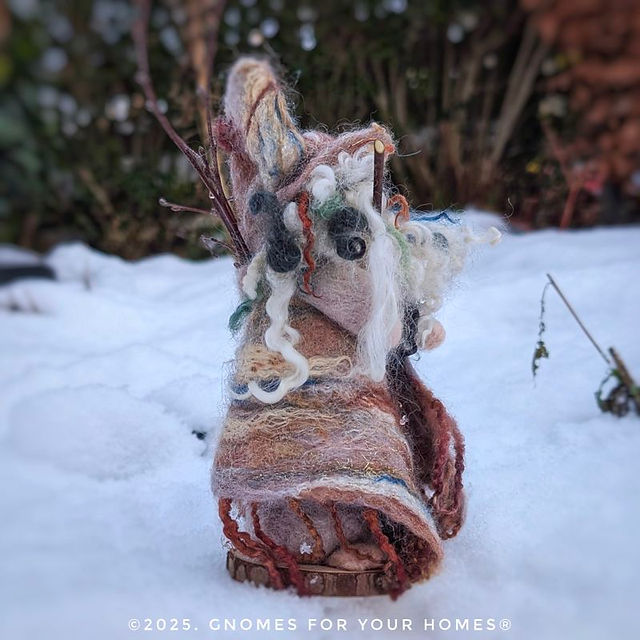 Alt text: A handcrafted felt figurine of a gnome or forest spirit called Beith standing in snow. The figure wears a textured brown and beige cloak made from felted wool, merino, and silk fibers. It has a distinctive facial features with wrinkles and a flowing white beard with curly locks. The character wears a pointed hat with Blue Faced Leicester wool curls and holds a wooden staff. The figurine's design includes subtle sparkly details and various natural textures, photographed against a snowy background. The image has a watermark "© 2025. GNOMES FOR YOUR HOMES®" at the bottom.