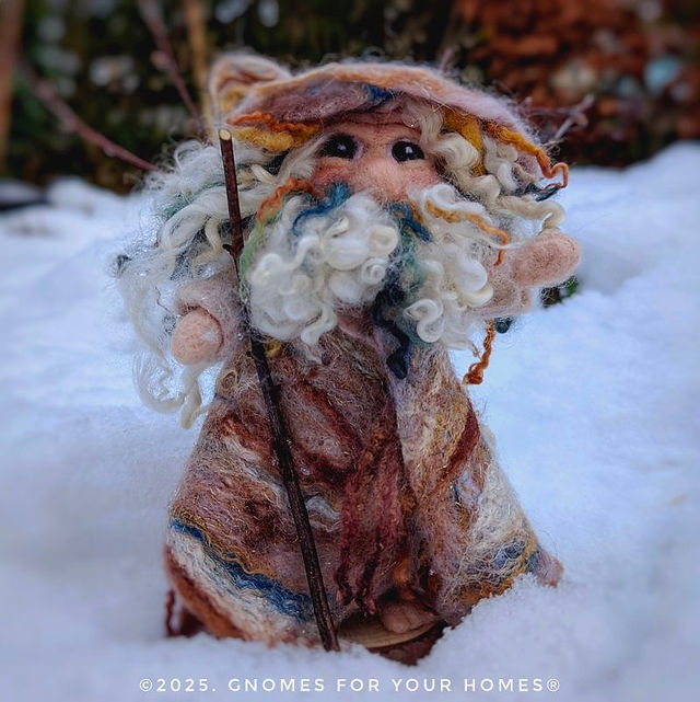 A handcrafted felt figurine of a gnome or forest spirit called Beith standing in snow. The figure wears a textured brown and beige cloak made from felted wool, merino, and silk fibers. It has a distinctive facial features with wrinkles and a flowing white beard with curly locks. The character wears a pointed hat with Blue Faced Leicester wool curls and holds a wooden staff. The figurine's design includes subtle sparkly details and various natural textures, photographed against a snowy background. The image has a watermark "© 2025. GNOMES FOR YOUR HOMES®" at the bottom.