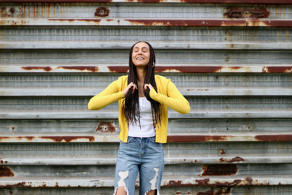 Woman in yellow cardigan combs braids in front of rusty wall