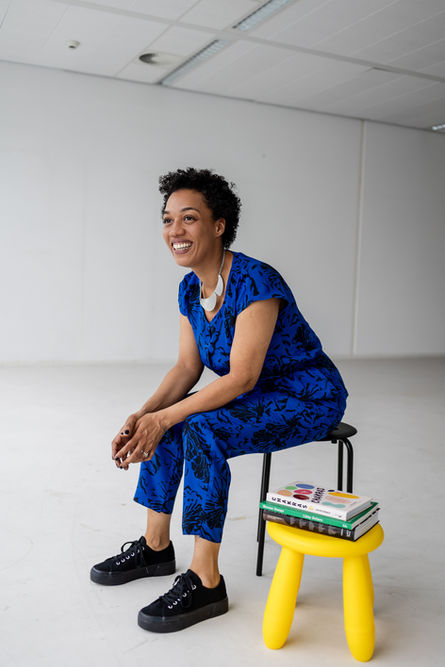 Black woman with short hair and blue jumpsuit sitting in studio with books and yellow table portrait photographer Brighton
