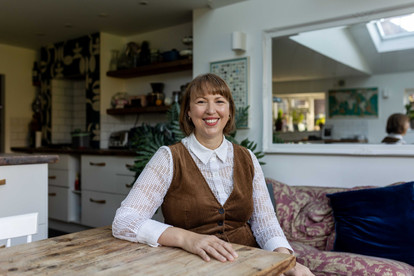 Smiling white woman in a white blouse and brown vest sits at a wooden table in a cozy kitchen. Plants and patterned walls in the background. Professional headshots Brighton