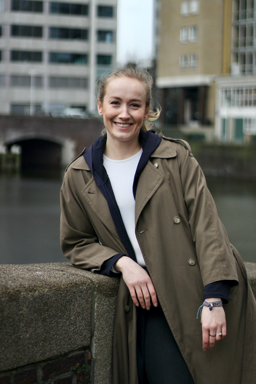 Blonde woman in brown coat leaning on stone wall in front of bridge