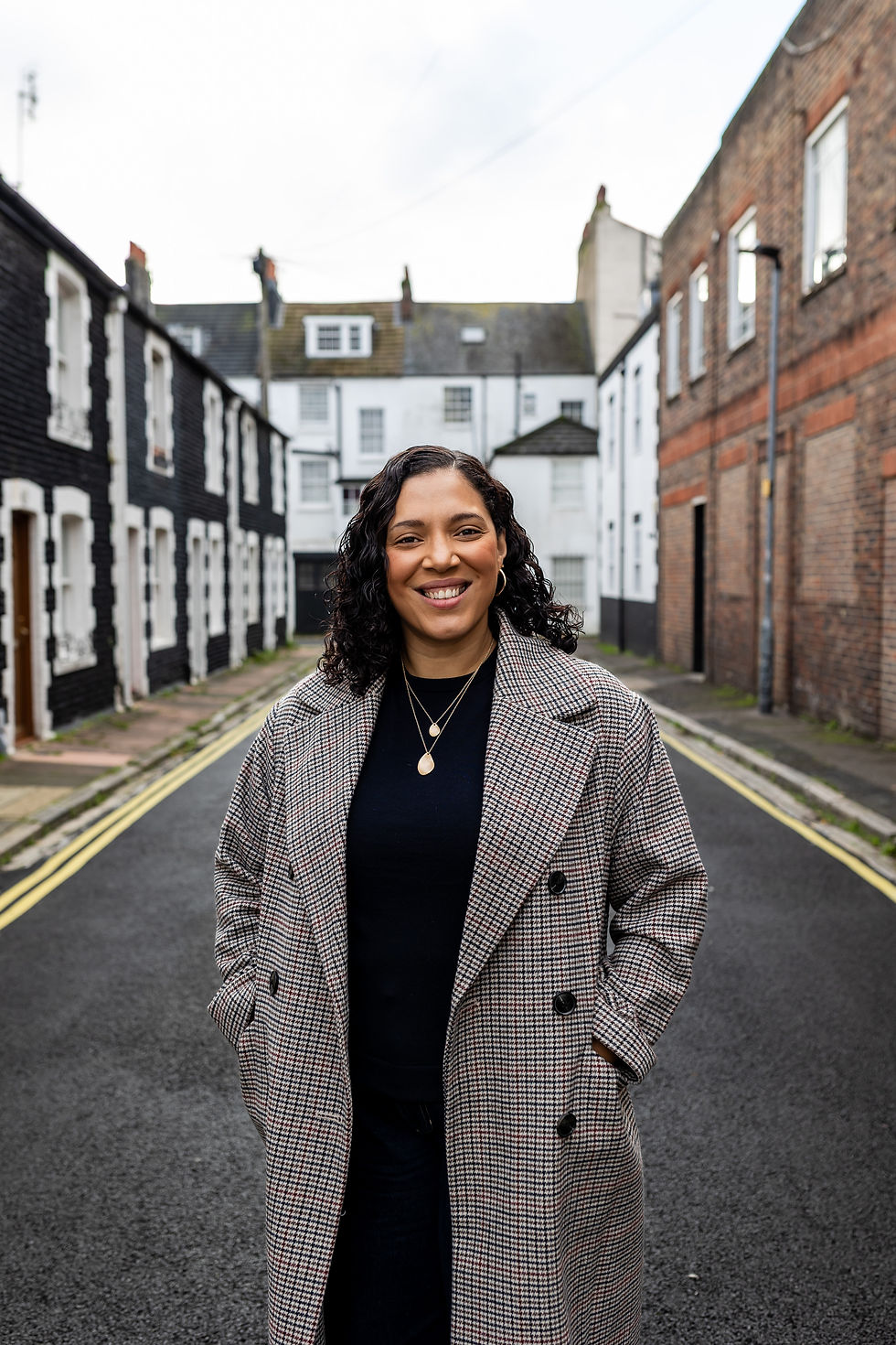 Woman with shoulder length curly black hair in grey checked jacket standing in the middle of a quiet street portrait photography Brighton
