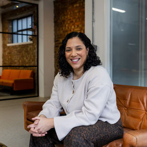 Woman with shoulder length black curly hair wearing beige cardigan and leopard print jeans sitting on orange leather sofa in open plan office portrait photography Brighton