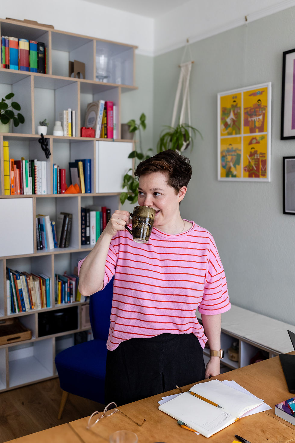 Creative personal branding white woman short brown hair pink tshirt drinking coffee at desk in front of colourful bookshelf
