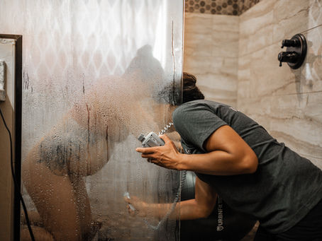 midwife in grey shit using stethoscope against a wet shower door while a birthing person labors in the shower
