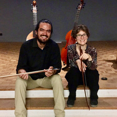 Jairo and Christine sit radiantly at the edge of the stage, each with a viol bow in their left hand. Behind them are two bass viols.