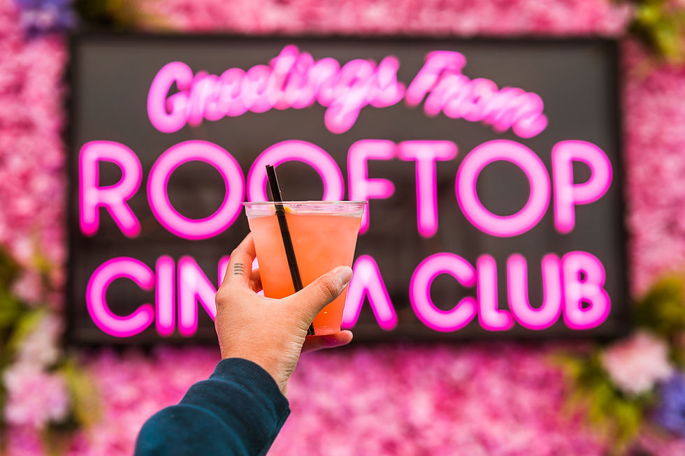 A hand holds a pink cocktail, with a neon RCC sign and decor in the background