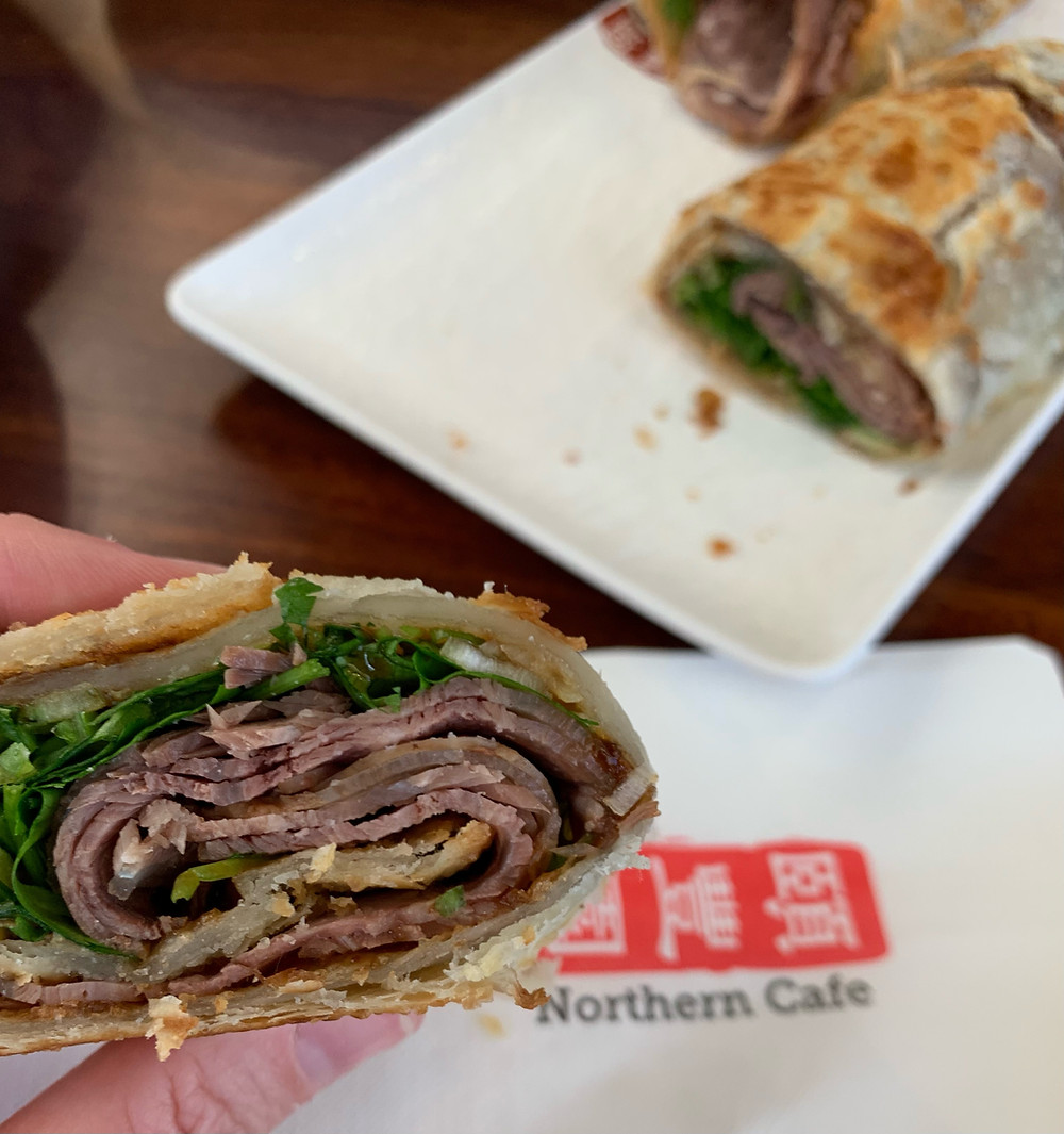 Marinated, thinly sliced beef coiled up with cilantro and green onion in a handmade pancake. Half of the roll is in a hand and half is on a plate. Northern Cafe napkin beneath
