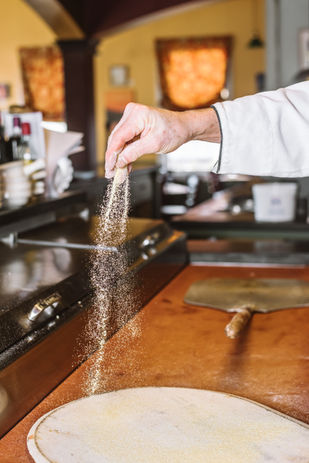 Chef sprinkling wooden pizza board to prevent dough sticking.