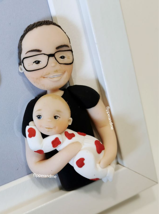 Statuina ritratto personalizzata di una donna con occhiali che tiene in braccio un neonato avvolto in una copertina bianca con cuori rossi, come sorpresa per annunciare una gravidanza.