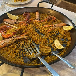 Paella dish on top of a restaurant table with a white tablecloth. The paella has langostines and rice and lemon and it has been mostly eaten. it has the serving spoon and fork resting on the paella dish.