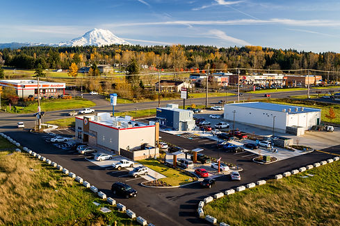 Aerial view of a suburban commercial retail center with parking lots and multiple buildings, with Mount Rainier visible in the background.