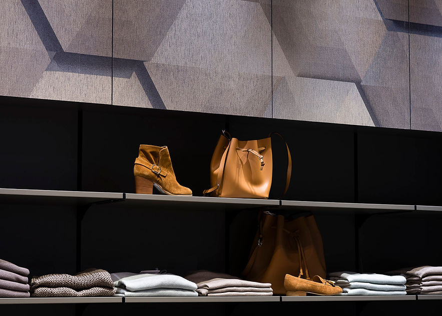 Detail eines Ladeninterieur in dunklen und gedeckten Farben. Im Vordergrund einige gelegte Kleidungsstücke, Schuhe und Taschen auf zwei eingelassenen Regalbrettern. Darüber ist die Wand mit einem grafischen Muster in Grautönen.
