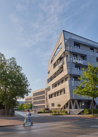 Teilansicht der markanten teiltransparenten Fassade des KPMG Gebäudes am Lenkwerk in Bielefeld. Im Vordergrund eine Strasse auf der eine Scooterfahrerin verwischt vorbeifährt. Auch die Fassaden der im Hintergrund zu sehenden Gebäude leuchten im Gegenlicht der Morgensonne.