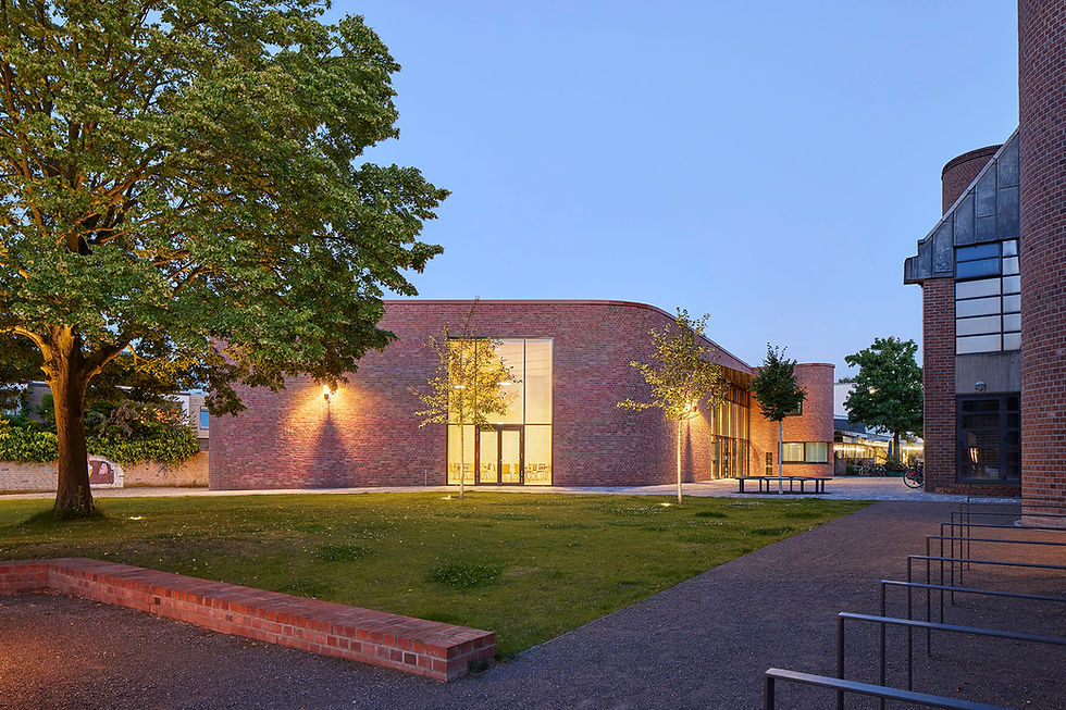 Blick über eine Wiese mit grünem Baum auf die beleuchtete rot geklinkerte Fassade der Nordseite des neuen Gemeindezentrum St. Matthäus im Abendlicht