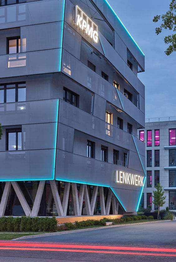 Detailansicht einer Ecke des KPMG-Gebäudes am Lenkwerk in Bielefeld. Der KPMG-Schriftzug und der Lenkwerk-Schriftzug sind weiss beleuchtet. Türkisfarbene Leuchten bilden mit der Fassade ein asymmetrisches Muster. Im Vordergrund eine Strasse mit verwischten Autorücklichtern.