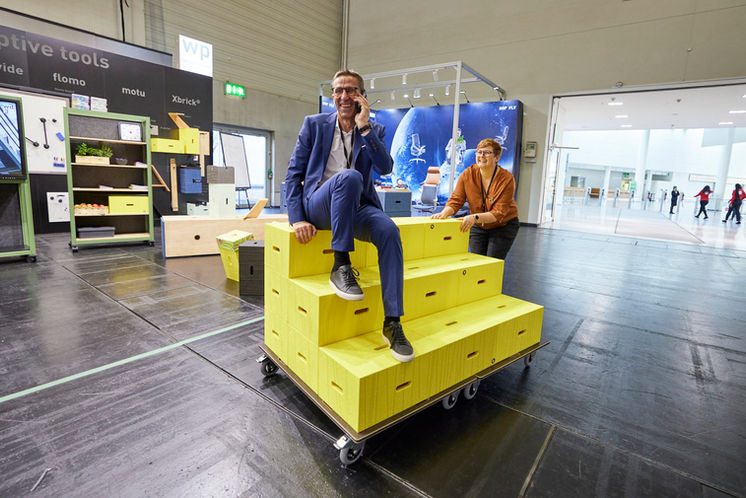 Ein telefonierender Mann in einem modernen blauen Anzug wird auf einem gelben Treppenelement mit Rollen von einer jungen Frau durch eine Messehalle gerollt, beide sind sehr fröhlich und lachen