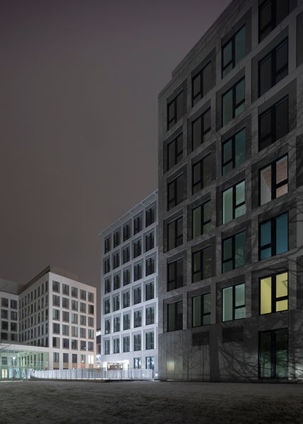 Nachtaufnahme von modernen Gebäuden mit grauen Fassaden und teilweise bunt erleuchteten Fenstern