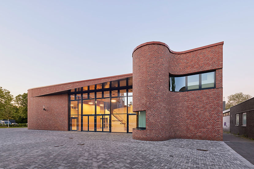 Blick über einen grau gepflasterten Platz auf den gesamten beleuchteten Eingangsbereich des neuen Gemeindezentrum St. Matthäus mit rundem Turm