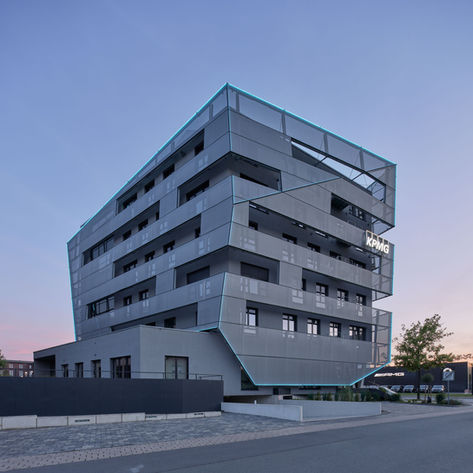 Gesamtansicht des KPMG Gebäudes am Lenkwerk in Bielefeld von der Ostseite im Licht der Dämmerung. Die Gebäudekanten werden durch türkisfarbene Leuchten akzentuiert. Der Himmel ist durch die gerade untergegangene Sonne blau und rot gefärbt. Im Vordergrund eine leere Strasse und Parkplätze.