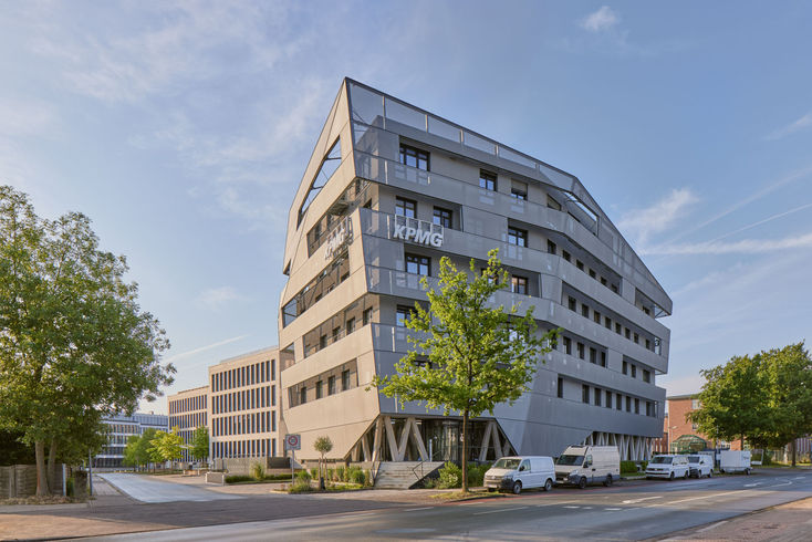 Das KPMG Gebäude am Lenkwerk in Bielefeld, davor steht ein grüner Baum. Im Vordergrund eine Strasse. Auch die Fassaden der im Hintergrund zu sehenden Gebäude leuchten im Gegenlicht der Morgensonne.