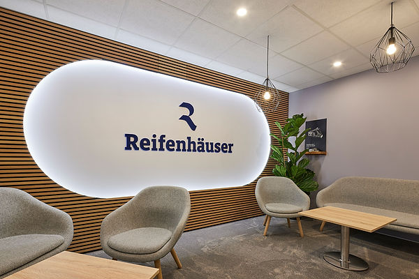 Edler Loungebereich mit Teppich und Sofa und Sesseln ohne Besucher, an der Wand der grosse Schriftzug REIFENHÄUSER