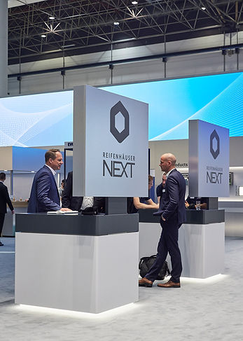 Besucher und Angestellte in Business-Kleidung unterhalten sich zwischen Präsentationstischen auf einem Messestand