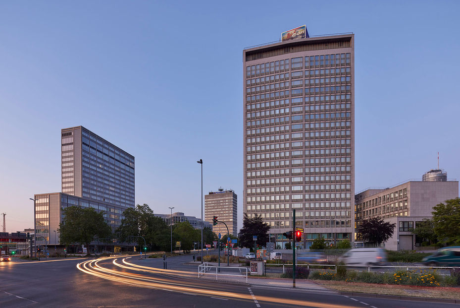 Fassade eines Hochhauses an einer großen stark befahrenen Kreuzung im Licht der Abenddämmerung, im Hintergrund und auf der linken Bildseite weitere große Gebäude.