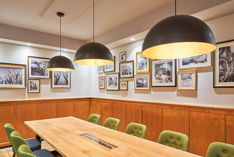 Blick über einen großen Holztisch mit 8 Stühlen über dem drei große runde Lampen hängen. An den Wänden hängen viele verschieden gerahmte Bilder, im unteren Bereich ist die Wand holzgetäfelt.
