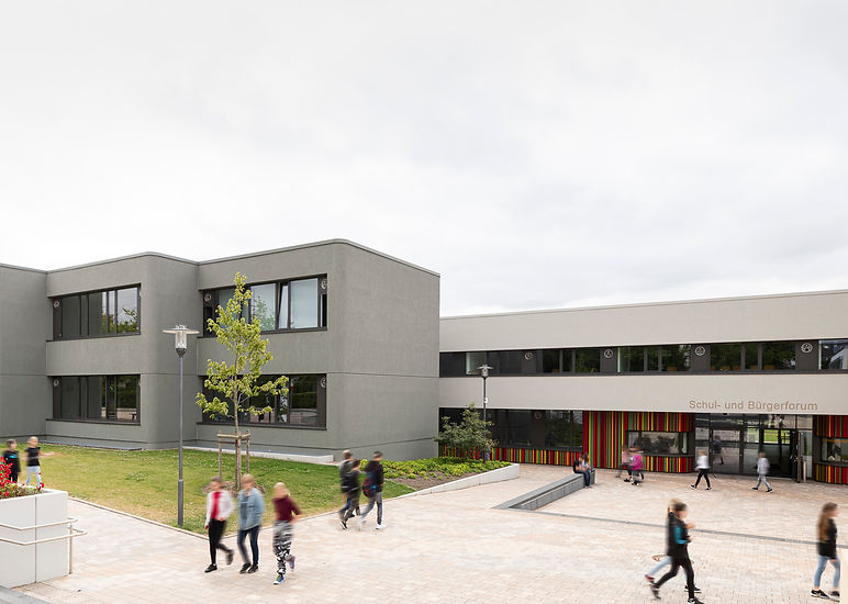 Frontalansicht des Eingangs des Schul- und Bürgerforum Barntrup mit einer Fassade aus bunten vertikalen Streifen im unteren Bereich, oben und links ist die Fassade aus Stein in grün-grau und beige-grau, im Vordergrund der Schulhof mit verwischten Schulkindern