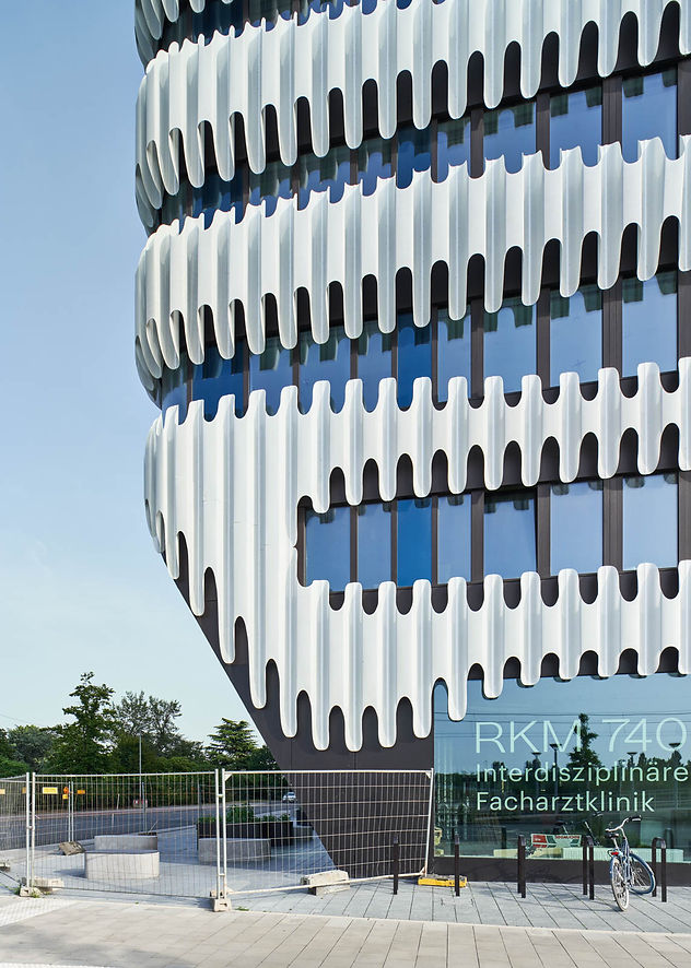 Gebäudeecke des RKM740 mit geschwungenen weissen Fassadenelementen und blauen Fenstern, im Vordergrund ein Fahrrad und ein Teil eines Bauzauns